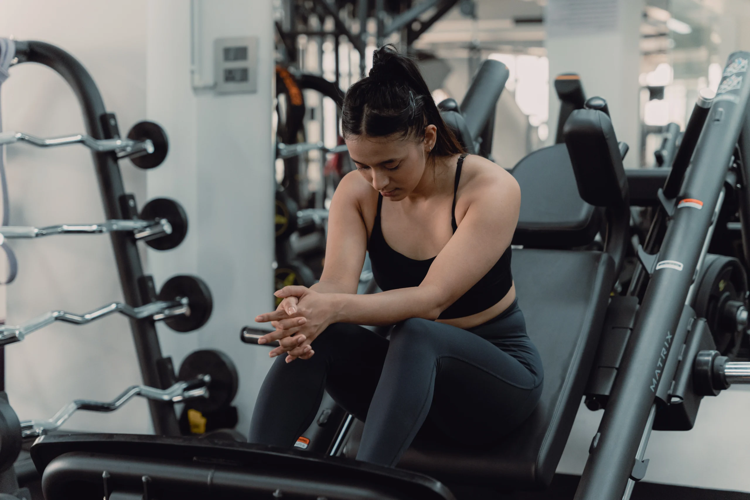 A young woman in athletic wear sits on a gym machine, looking down with her hands clasped together. She appears deep in thought, possibly feeling gym anxiety.