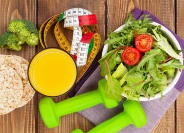 Best Foods to Eat Before and After a Workout, featuring a healthy salad with tomatoes, fresh broccoli, rice cakes, orange juice, and dumbbells on a wooden table.