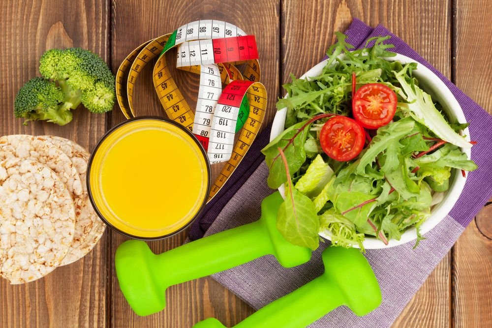 Best Foods to Eat Before and After a Workout, featuring a healthy salad with tomatoes, fresh broccoli, rice cakes, orange juice, and dumbbells on a wooden table.